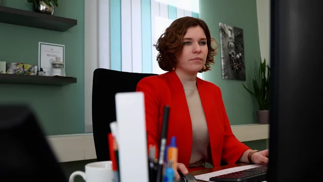 Eine konzentrierte Frau mit lockigem Haar sitzt an einem Schreibtisch und arbeitet am Computer. Sie trägt eine rote Jacke über einem hellen Oberteil. Im Hintergrund sind Pflanzen und einige Gegenstände auf einem Regal sichtbar.