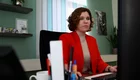 Eine konzentrierte Frau mit lockigem Haar sitzt an einem Schreibtisch und arbeitet am Computer. Sie trägt eine rote Jacke über einem hellen Oberteil. Im Hintergrund sind Pflanzen und einige Gegenstände auf einem Regal sichtbar.