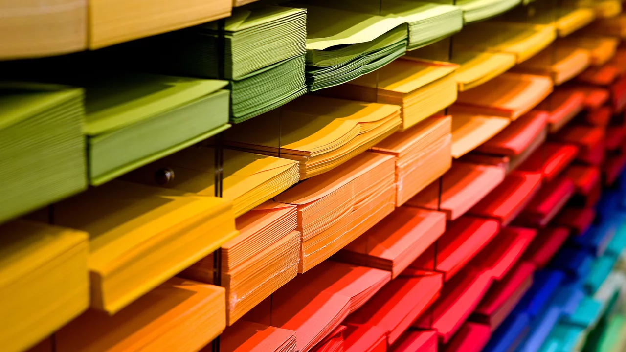 Ein Regal mit farbigem Papier in verschiedenen Tönen. Die Farben verlaufen von grün über gelb, orange und rot bis hin zu blau. Die Papiere sind ordentlich gestapelt und zeigen eine harmonische Farbverlauf.