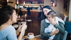 Zwei Frauen sitzen in einem Café. Eine Frau hält ein Baby in den Armen und lächelt die andere an, die ihr gegenüber sitzt. Auf dem Tisch steht eine Tasse Kaffee. Die Atmosphäre ist freundlich und entspannt.