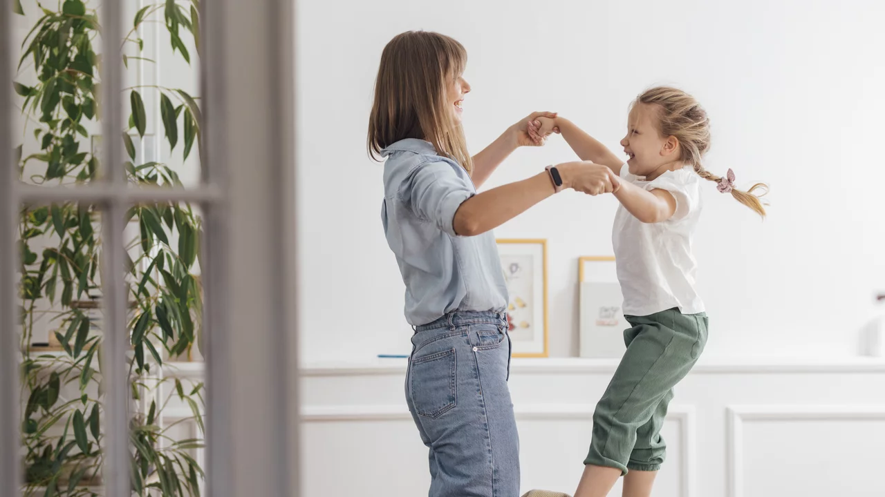 Eine Mutter tanzt mit ihrer Tochter auf dem Bett. Die Tochter steht auf dem Bett, lachend und fröhlich, während die Mutter ihre Hände hält. Der Raum ist hell eingerichtet, mit Pflanzen im Hintergrund.