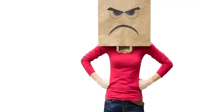 Eine Person trägt eine bemalte Papiertüte als Kopf, die ein wütendes Gesicht zeigt. Sie steht mit verschränkten Armen da und trägt ein rotes Oberteil. Die Körpersprache drückt Unzufriedenheit oder Ärger aus.