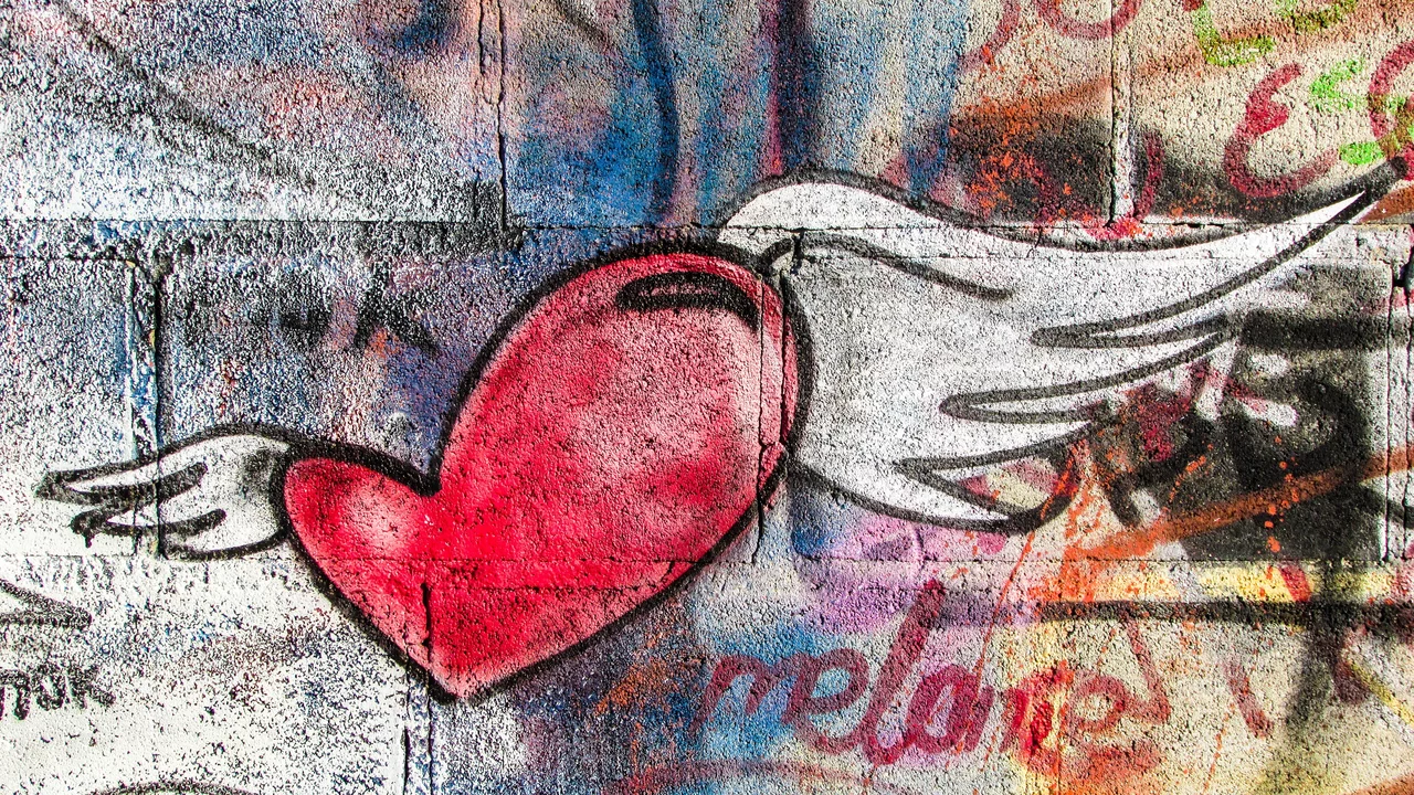 Eine stilisierte Darstellung eines roten Herzens mit weißen Flügeln, das in einem bunten Graffiti-Hintergrund eingebettet ist. Das Bild vermittelt ein Gefühl von Freiheit und Liebe.