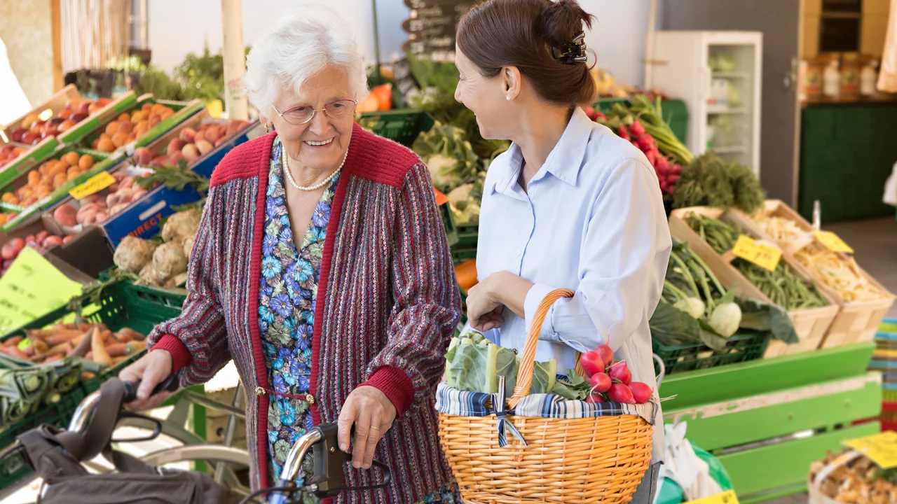 Eine ältere Frau mit einem Rollator steht an einem Marktstand mit frischem Obst und Gemüse. Sie lächelt eine jüngere Frau an, die einen Korb mit Gemüse in der Hand hält. Beide wirken freundlich und genießen den Moment beim Einkaufen.