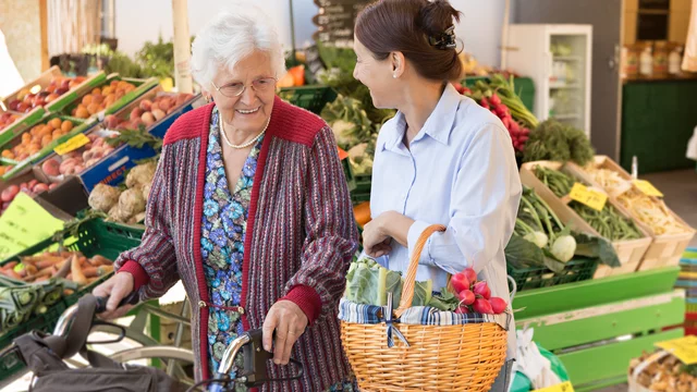 Eine ältere Frau mit einem Rollator steht an einem Marktstand mit frischem Obst und Gemüse. Sie lächelt eine jüngere Frau an, die einen Korb mit Gemüse in der Hand hält. Beide wirken freundlich und genießen den Moment beim Einkaufen.