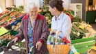 Eine ältere Frau mit einem Rollator steht an einem Marktstand mit frischem Obst und Gemüse. Sie lächelt eine jüngere Frau an, die einen Korb mit Gemüse in der Hand hält. Beide wirken freundlich und genießen den Moment beim Einkaufen.