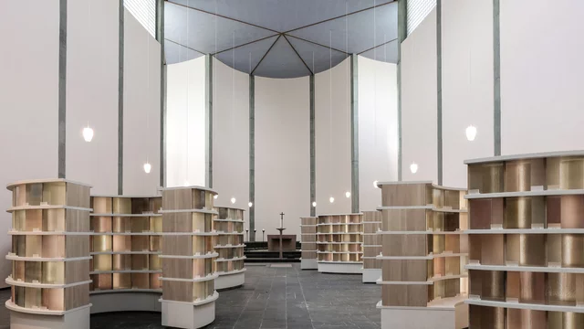 Die Darstellung zeigt einen modernen Raum mit mehreren Buchregalen, die in geschwungener Anordnung angeordnet sind. Der Boden ist aus dunklem Stein, und die Wände sind hoch und hell. In der Mitte steht ein Tisch, und es gibt eine sanfte Beleuchtung von hängenden Lampen.