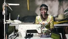 Eine Frau mit geflochtenen Haaren sitzt an einer Nähmaschine in einem Raum mit vielen Stoffen und Materialien. Sie trägt ein buntes Kleid und lächelt freundlich in die Kamera, während sie an einem Nähprojekt arbeitet.