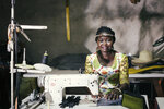 Eine Frau mit geflochtenen Haaren sitzt an einer Nähmaschine in einem Raum mit vielen Stoffen und Materialien. Sie trägt ein buntes Kleid und lächelt freundlich in die Kamera, während sie an einem Nähprojekt arbeitet.