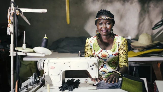 Eine Frau mit geflochtenen Haaren sitzt an einer Nähmaschine in einem Raum mit vielen Stoffen und Materialien. Sie trägt ein buntes Kleid und lächelt freundlich in die Kamera, während sie an einem Nähprojekt arbeitet.