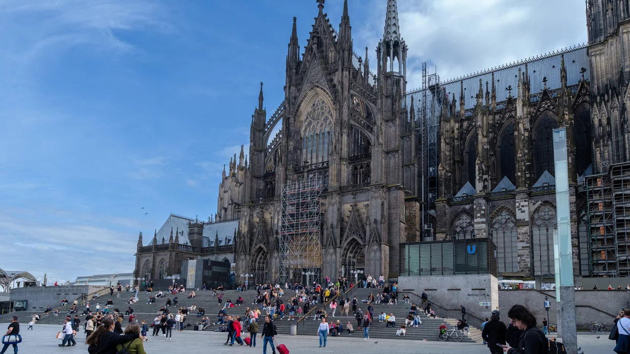 Ein großer gotischer Kathedrale mit vielen Türmen und Fenster im Hintergrund. Vor der Kathedrale sind zahlreiche Menschen zu sehen, die auf Stufen und im Freien sitzen. Der Himmel ist teilweise bewölkt, und einige Teile des Gebäudes sind eingerüstet.