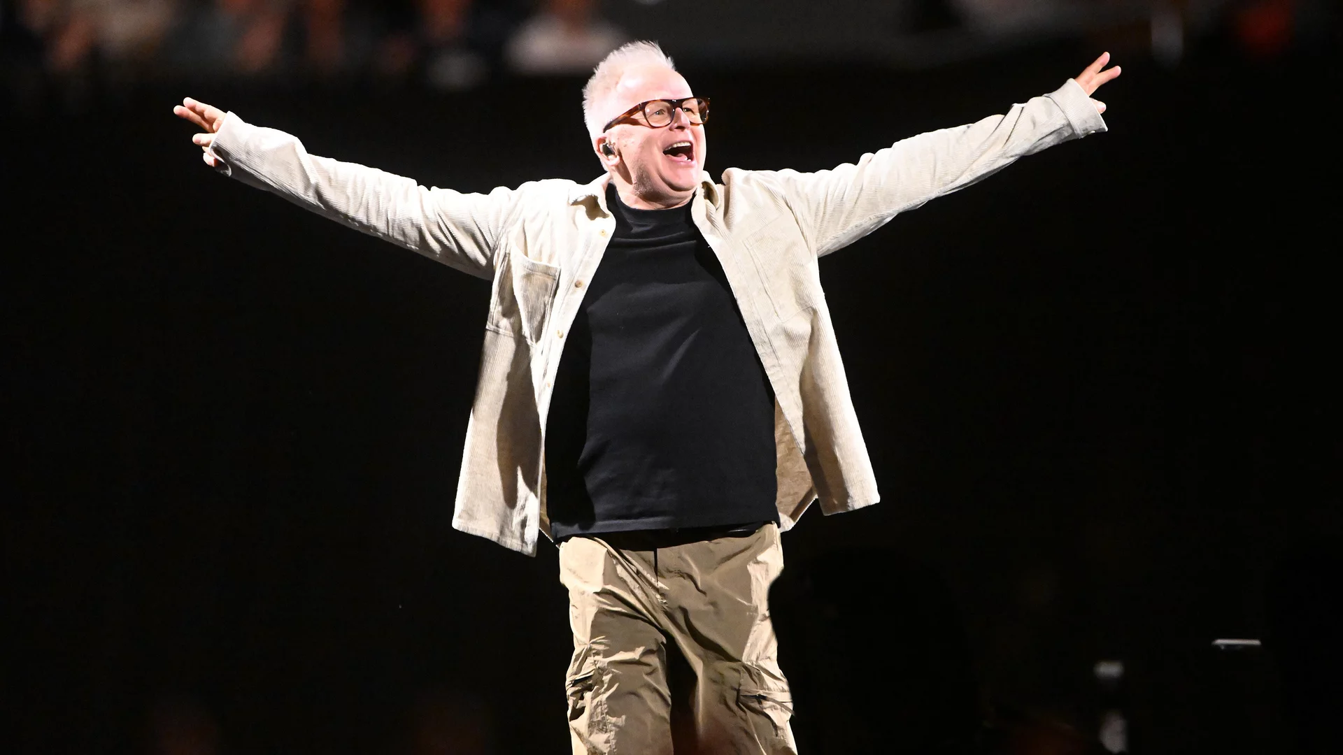 Ein enthusiastischer Mann mit kurzen, grauen Haaren und Brille steht mit ausgebreiteten Armen auf einer Bühne. Er trägt ein schwarzes T-Shirt und eine helle Jacke, umgeben von einer dunklen Kulisse und einer jubelnden Menge im Hintergrund.