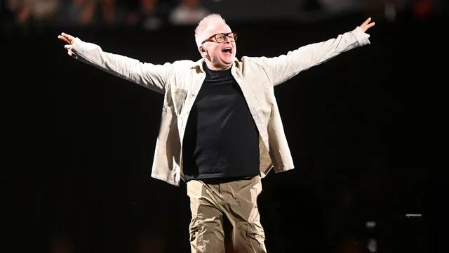 Ein enthusiastischer Mann mit kurzen, grauen Haaren und Brille steht mit ausgebreiteten Armen auf einer Bühne. Er trägt ein schwarzes T-Shirt und eine helle Jacke, umgeben von einer dunklen Kulisse und einer jubelnden Menge im Hintergrund.