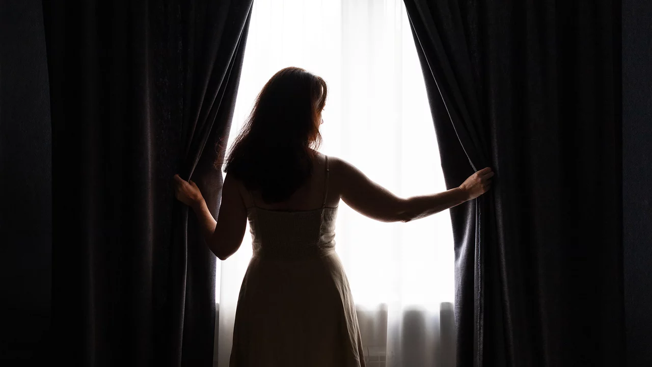 Eine Frau steht mit dem Rücken zur Kamera und zieht schweres Vorhänge auf. Im Hintergrund ist ein helles Fenster zu sehen, durch das Licht strömt. Die Szene vermittelt ein Gefühl von Erwartung und Neuanfang.