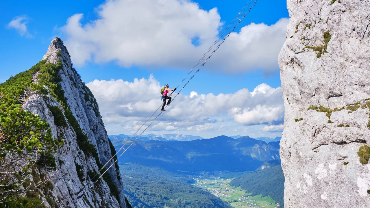 Eine Person klettert an einer steilen Felswand und nutzt dabei ein Seil oder eine Leiternstruktur. Im Hintergrund erstrecken sich Berge und ein Tal unter einem blauen Himmel mit einigen Wolken. Die Szene vermittelt ein Gefühl von Abenteuer und Hochgebirgen.