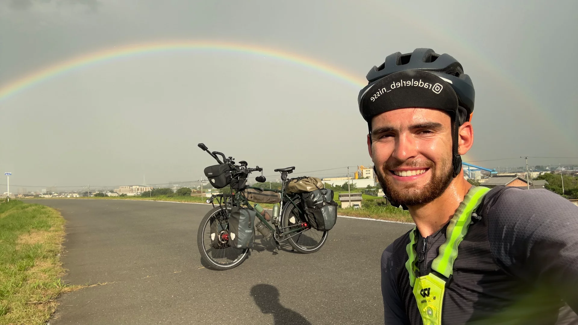 Ein junger Mann mit Helm lächelt vor einer Straße. Im Hintergrund steht ein beladenes Fahrrad und ein Regenbogen ist am Himmel sichtbar.