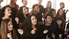 Eine Gruppe von jungen Menschen singt und bewegt sich gemeinsam in einem Raum. Sie tragen schwarze Kleidung und zeigen Freude und Engagement beim Singen. Die Szene strahlt Gemeinschaft und Begeisterung aus.