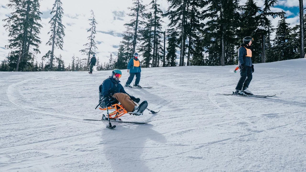 Eine Gruppe von Skifahrern auf einer verschneiten Piste. Ein Skifahrer sitzt auf einem speziellen Schlitten, während andere stehend fahren. Hohe Tannenbäume umgeben die Szene, und der Himmel ist teilweise bewölkt.
