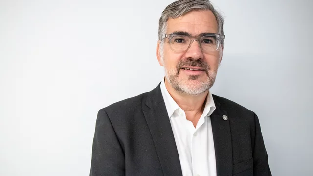 Ein Mann mit Brille, grau melierten Haaren und einem Bart trägt einen schwarzen Anzug und ein weißes Hemd. Er steht vor einer hellen Wand und lächelt freundlich, was eine professionelle und einladende Ausstrahlung vermittelt.