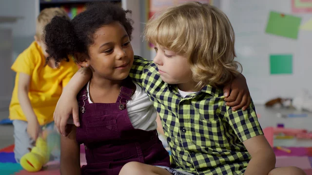 Zwei Kinder sitzen eng beieinander. Ein Mädchen mit lockigem Haar trägt ein lila Latzkleid und schaut lächelnd zu einem Jungen mit kurzen blonden Haaren, der kariertes Shirt trägt. Sie scheinen Spaß zu haben und eine freundliche Verbindung zu teilen.