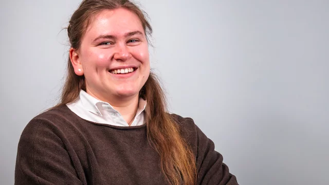 Eine lächelnde junge Frau mit langen Haaren trägt einen braunen Pullover und ein weißes Hemd. Sie sitzt mit verschränkten Armen vor einem hellen Hintergrund und strahlt Freundlichkeit und Selbstsicherheit aus.