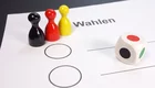 Auf dem Bild sind drei Spielsteine in Schwarz, Rot und Gelb sowie ein Würfel abgebildet. Vor ihnen liegt ein Wahlzettel mit zwei leeren Kästchen und dem Wort „Wahlen“. Das Bild vermittelt das Thema der Auswahl oder Abstimmung.