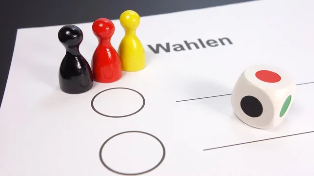 Auf dem Bild sind drei Spielsteine in Schwarz, Rot und Gelb sowie ein Würfel abgebildet. Vor ihnen liegt ein Wahlzettel mit zwei leeren Kästchen und dem Wort „Wahlen“. Das Bild vermittelt das Thema der Auswahl oder Abstimmung.