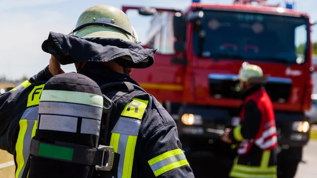 Ein Feuerwehrmann in Schutzausrüstung steht mit dem Rücken zur Kamera, während ein rotes Feuerwehrauto im Hintergrund parkt. Ein weiterer Feuerwehrmann in roter Weste ist ebenfalls sichtbar. Es scheint eine Einsatzsituation zu sein, in der die Feuerwehr aktiv ist.