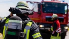 Ein Feuerwehrmann in Schutzausrüstung steht mit dem Rücken zur Kamera, während ein rotes Feuerwehrauto im Hintergrund parkt. Ein weiterer Feuerwehrmann in roter Weste ist ebenfalls sichtbar. Es scheint eine Einsatzsituation zu sein, in der die Feuerwehr aktiv ist.