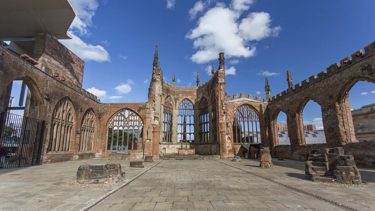 Eine weitläufige Ansicht der Ruinen einer historischen Kirche mit gotischen Fenstern und hohen Mauern unter einem klaren blauen Himmel. Der Boden ist gepflastert, und einige alte Steinstücke sind sichtbar. Die Szene vermittelt einen Eindruck von Geschichte und Erhabenheit.