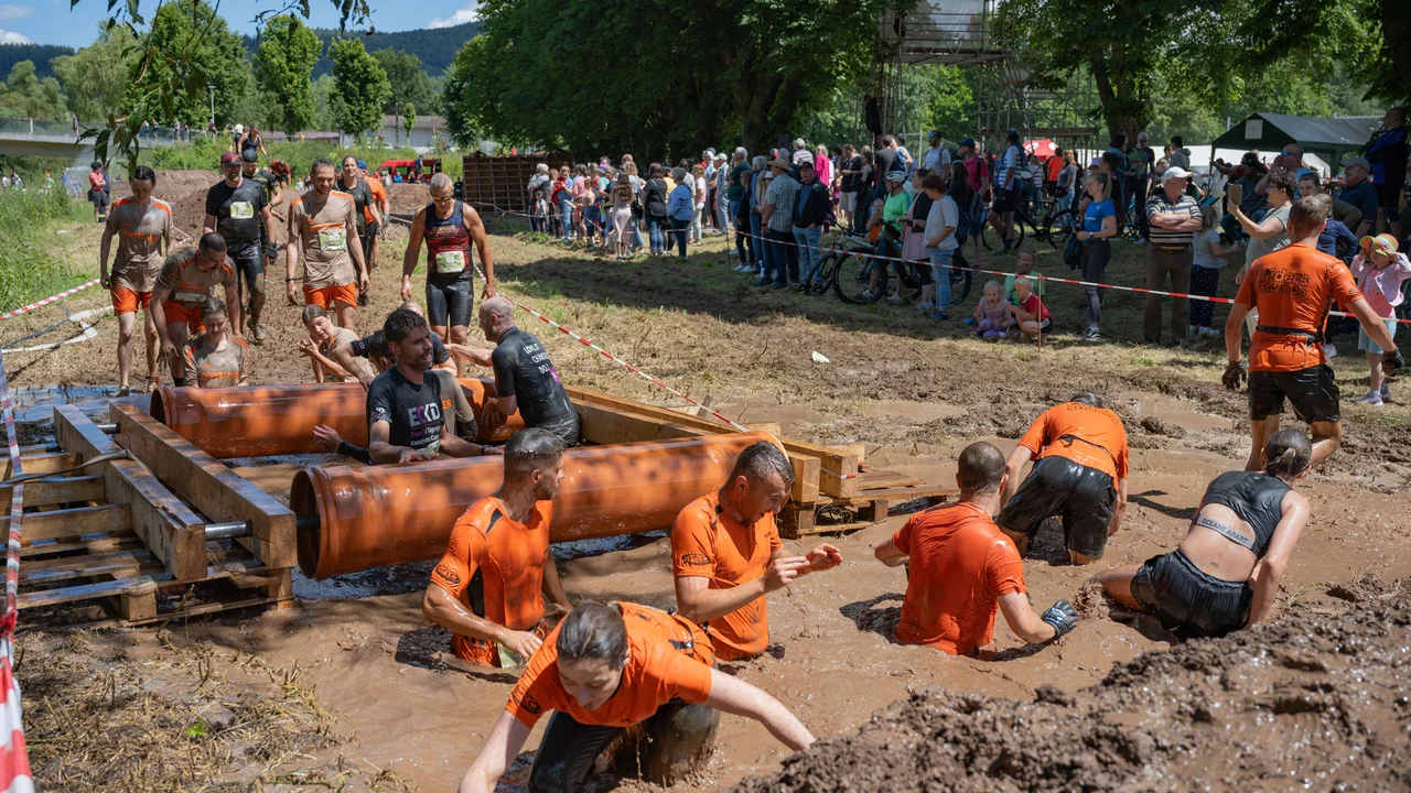 Eine Gruppe von Teilnehmern in orangefarbener Sportkleidung kämpft sich durch einen schmutzigen Hindernisparcours mit Wasser. Einige schwimmen im Matsch, während andere sich auf Holzplanken abstützen. Im Hintergrund beobachten zahlreiche Zuschauer die Veranstaltung.