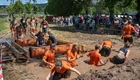 Eine Gruppe von Teilnehmern in orangefarbener Sportkleidung kämpft sich durch einen schmutzigen Hindernisparcours mit Wasser. Einige schwimmen im Matsch, während andere sich auf Holzplanken abstützen. Im Hintergrund beobachten zahlreiche Zuschauer die Veranstaltung.