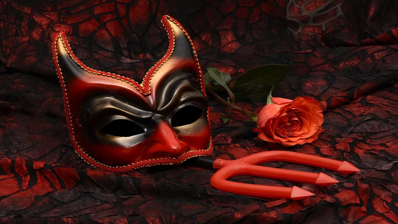 Ein rotes Teufelsmaske mit Hörnern liegt auf einem dunklen, strukturierten Untergrund. Neben der Maske befinden sich eine rote Gabel und eine rosa Rose. Die Farben wirken intensiv und schaffen eine geheimnisvolle Atmosphäre.