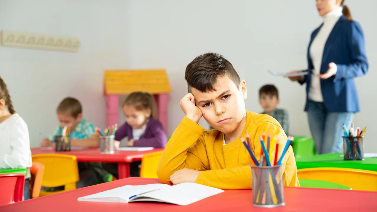 Blick in ein Klassenzimmer. Am Tisch im Vordergrund sitzt ein Junge, den Kopf in seine rechte Hand gestützt und schmollt. Im Hintergrund sieht man die Lehrerin, die den anderen Kindern etwas erklärt.