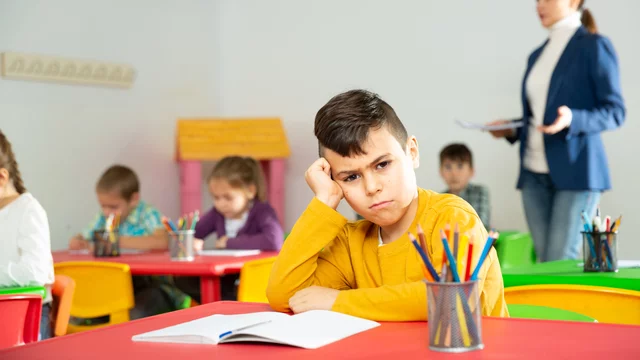 Blick in ein Klassenzimmer. Am Tisch im Vordergrund sitzt ein Junge, den Kopf in seine rechte Hand gestützt und schmollt. Im Hintergrund sieht man die Lehrerin, die den anderen Kindern etwas erklärt.