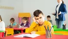 Blick in ein Klassenzimmer. Am Tisch im Vordergrund sitzt ein Junge, den Kopf in seine rechte Hand gestützt und schmollt. Im Hintergrund sieht man die Lehrerin, die den anderen Kindern etwas erklärt.