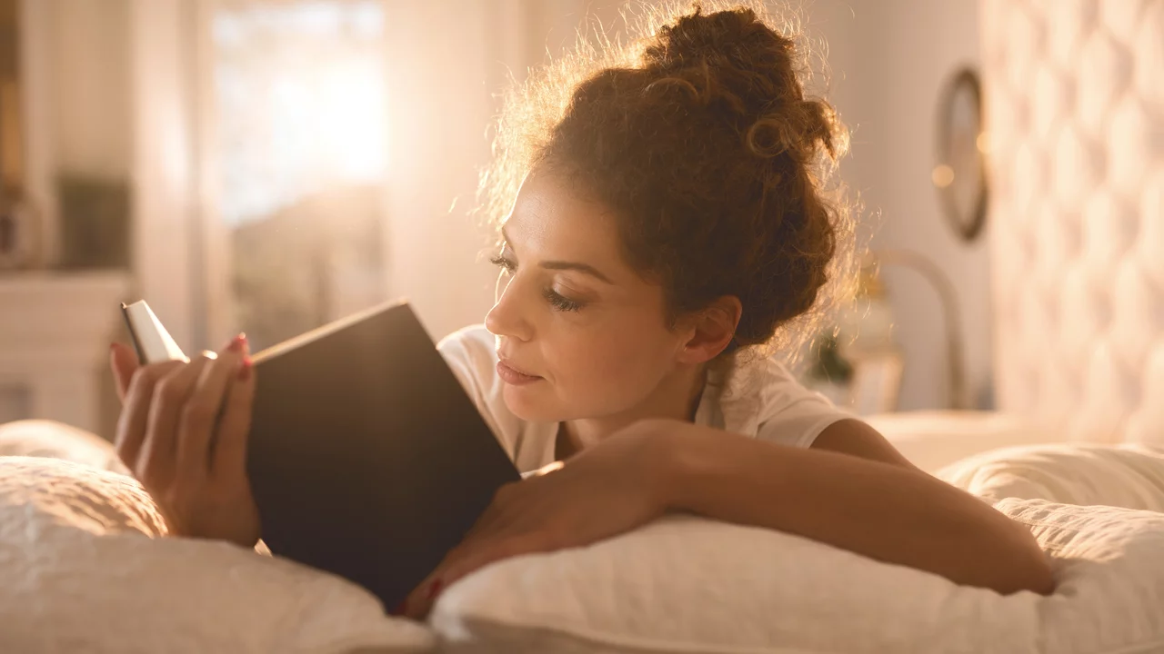 Eine junge Frau liegt auf einem Bett und liest ein Buch. Sie hat lockiges, braunes Haar und ist in einem entspannten Moment vertieft. Sanftes Licht durchströmt den Raum und schafft eine gemütliche Atmosphäre.