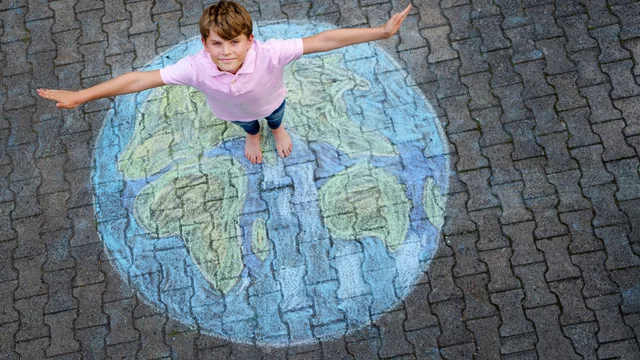 Ein Junge steht barfuß auf einem Gehweg, seine Arme ausgebreitet. Unter ihm ist eine bunte, kreidegemalte Weltkugel zu sehen, die den Kontinenten darstellt. Der Junge trägt ein pinkes Poloshirt und lächelt.