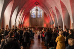 Eine große Gruppe von Menschen steht in einer kirchlichen Umgebung. Im Vordergrund sind zwei Personen in liturgischen Gewändern zu sehen, während im Hintergrund Fenster das Licht farblich verbreiten. Die Stimmung ist feierlich und andächtig, und die Menschen scheinen an einem Gottesdienst teilzunehmen.