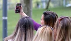 Eine Gruppe von vier Frauen steht zusammen und macht ein Selfie mit einem Smartphone. Die Frau, die das Foto aufnimmt, lächelt, während die anderen in die Kamera schauen. Es ist ein fröhlicher Moment im Freien.