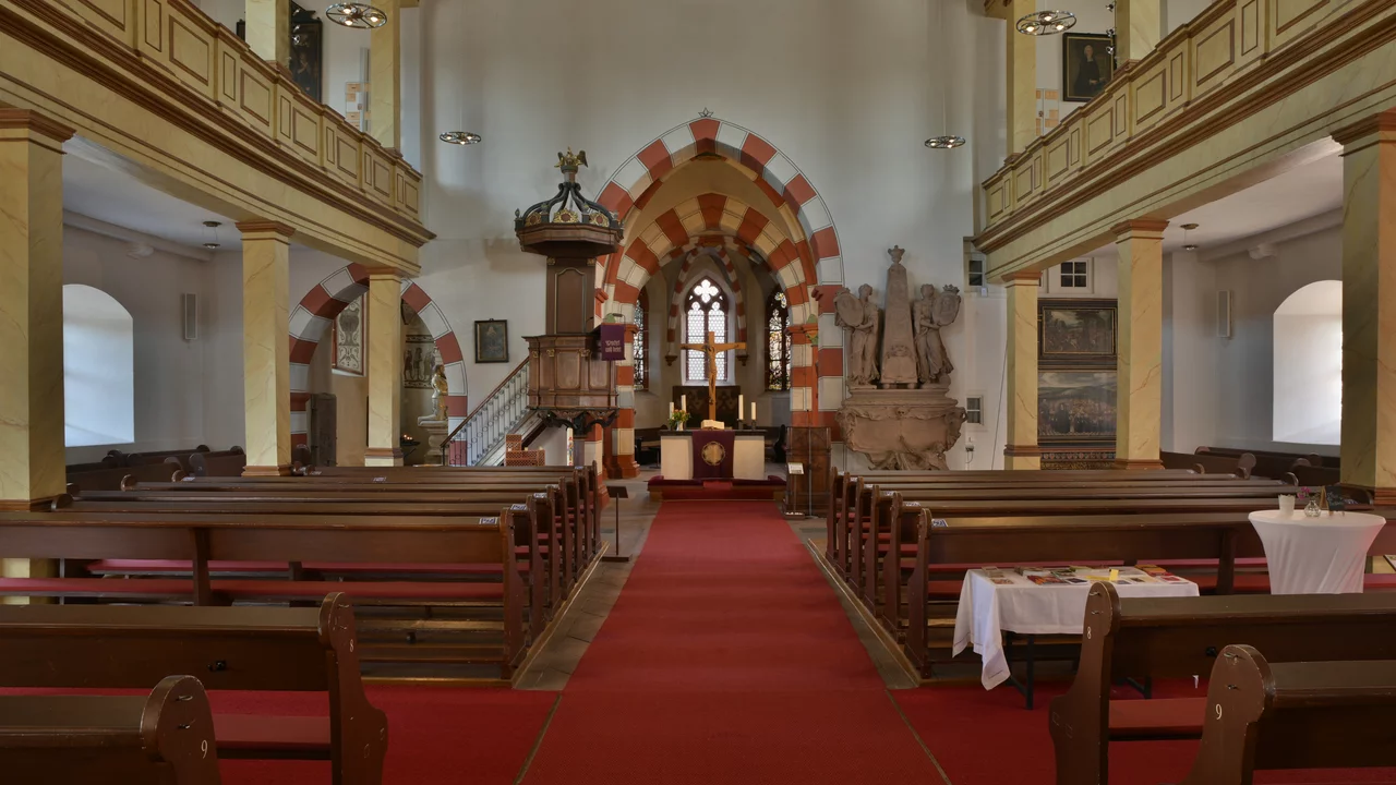 Eine Ansicht des Innenraums einer Kirche mit Holzbankreihen und einem roten Teppich in der Mitte. An der Wand sind bunte Fenster und ein Haupaltar sichtbar. Eine Kanzel steht links im Raum, während rechts eine Statue zu sehen ist. Der Raum wirkt hell und einladend.