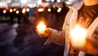 Eine Person hält zwei brennende Kerzen in der Hand und steht in einer Menschenmenge. Das Licht der Kerzen strahlt leicht und sorgt für eine feierliche Atmosphäre in der Umgebung.