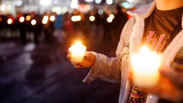 Eine Person hält zwei brennende Kerzen in der Hand und steht in einer Menschenmenge. Das Licht der Kerzen strahlt leicht und sorgt für eine feierliche Atmosphäre in der Umgebung.