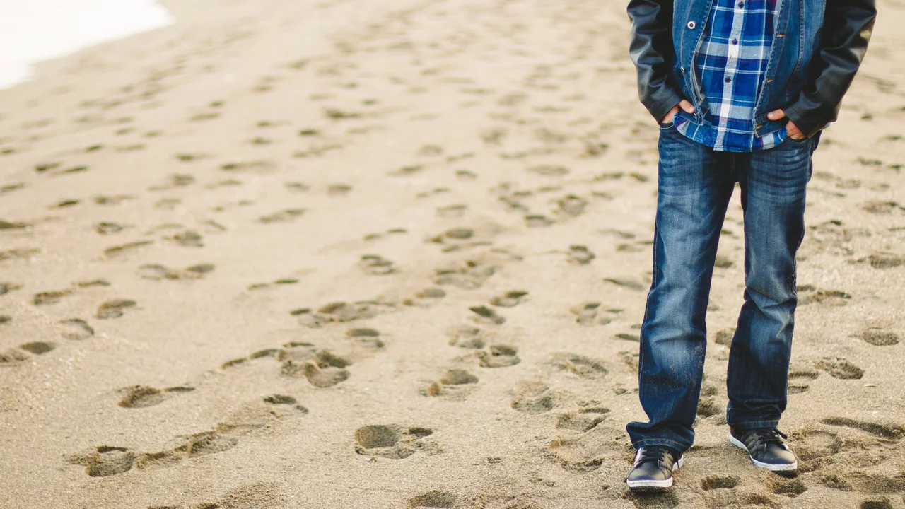 Ein Junge steht am Strand, mit den Händen in den Taschen, bekleidet mit einer karierten Hemd und Jeans. Sand und Fußabdrücke sind im Vordergrund sichtbar.