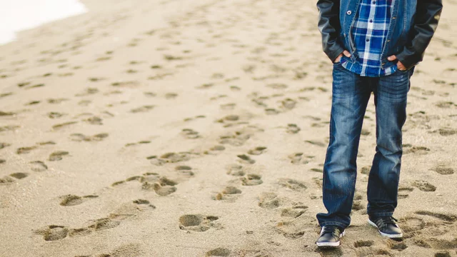 Ein Junge steht am Strand, mit den Händen in den Taschen, bekleidet mit einer karierten Hemd und Jeans. Sand und Fußabdrücke sind im Vordergrund sichtbar.