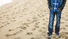 Ein Junge steht am Strand, mit den Händen in den Taschen, bekleidet mit einer karierten Hemd und Jeans. Sand und Fußabdrücke sind im Vordergrund sichtbar.