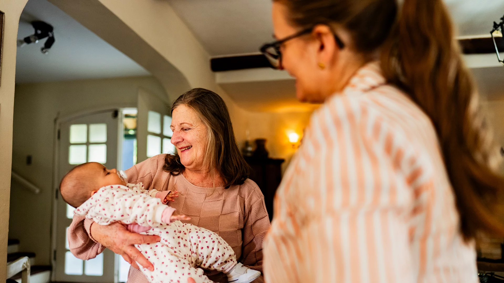 Eine ältere Frau hält ein Baby in den Armen und lächelt. Neben ihr steht eine Frau in einem gestreiften Oberteil, die ebenfalls lächelt. Die Szene vermittelt eine warme, familiäre Atmosphäre.
