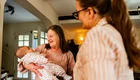 Eine ältere Frau hält ein Baby in den Armen und lächelt. Neben ihr steht eine Frau in einem gestreiften Oberteil, die ebenfalls lächelt. Die Szene vermittelt eine warme, familiäre Atmosphäre.