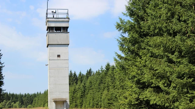 Ein betonsäulenförmiger Beobachtungsturm steht in einer offenen Wiese, umgeben von einer dichten Baumreihe. Der Himmel ist überwiegend blau mit einigen Wolken. Der Turm hat eine kleine Plattform oben, wo sich offenbar eine Überwachungskamera befindet.