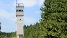 Ein betonsäulenförmiger Beobachtungsturm steht in einer offenen Wiese, umgeben von einer dichten Baumreihe. Der Himmel ist überwiegend blau mit einigen Wolken. Der Turm hat eine kleine Plattform oben, wo sich offenbar eine Überwachungskamera befindet.
